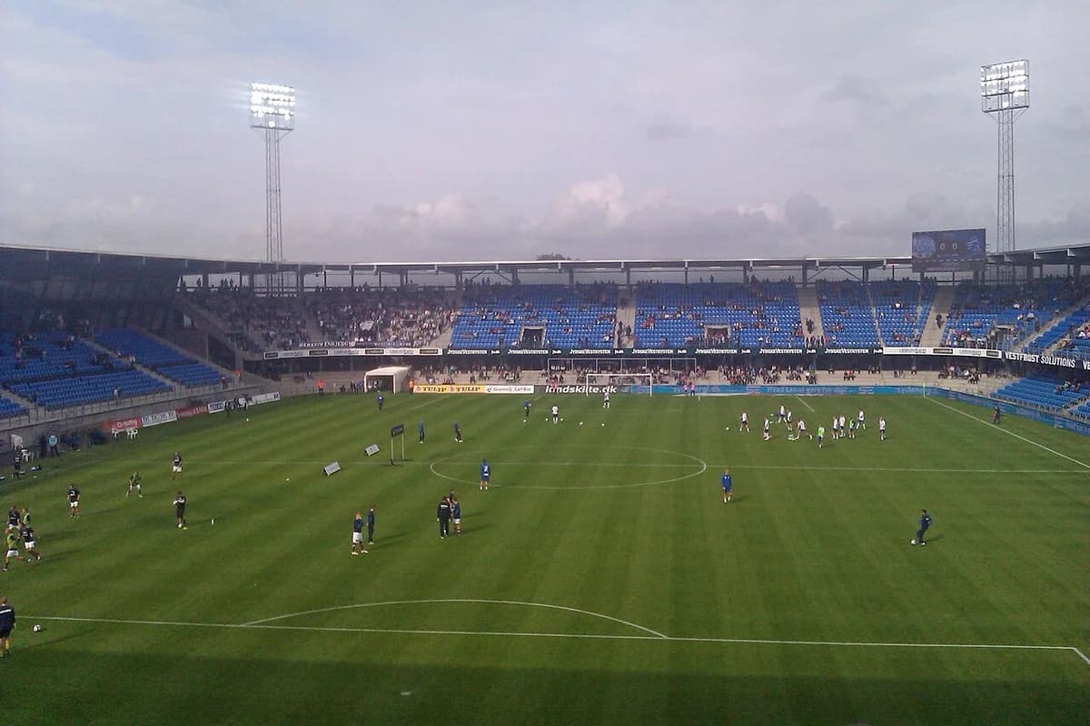 Football players warming up on the pitch at a stadium with blue seats and floodlights.