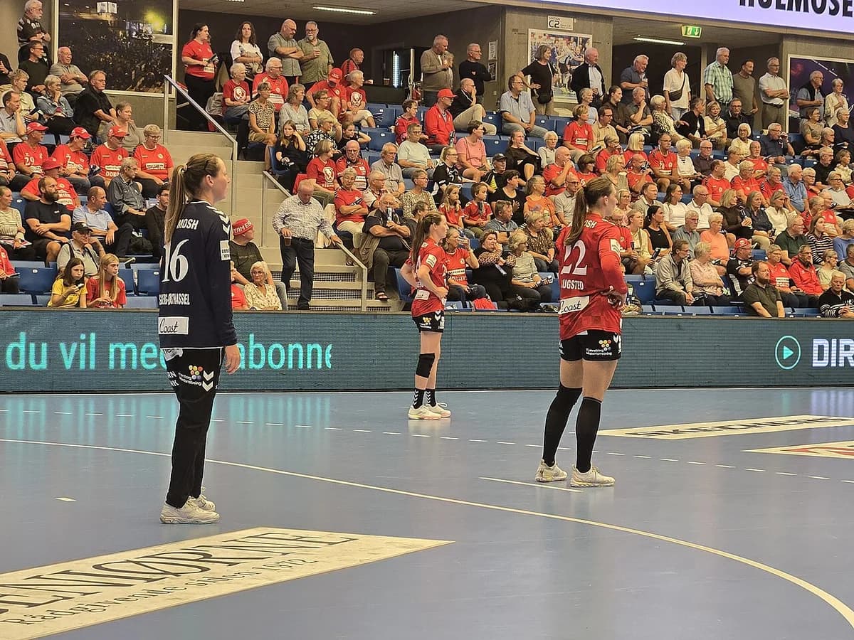 Women’s handball game with players on court and a crowded stand in the background.