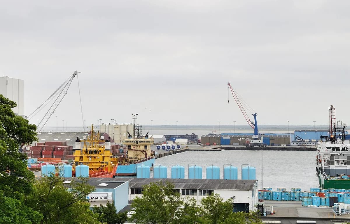Industrial harbor scene with cranes, ships, and containers seen across the water.